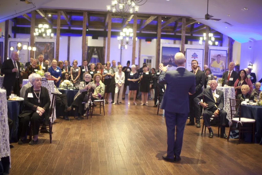 A speaker addressing a large group of guests seated and standing at a formal fundraising event at Stevenson Ridge.