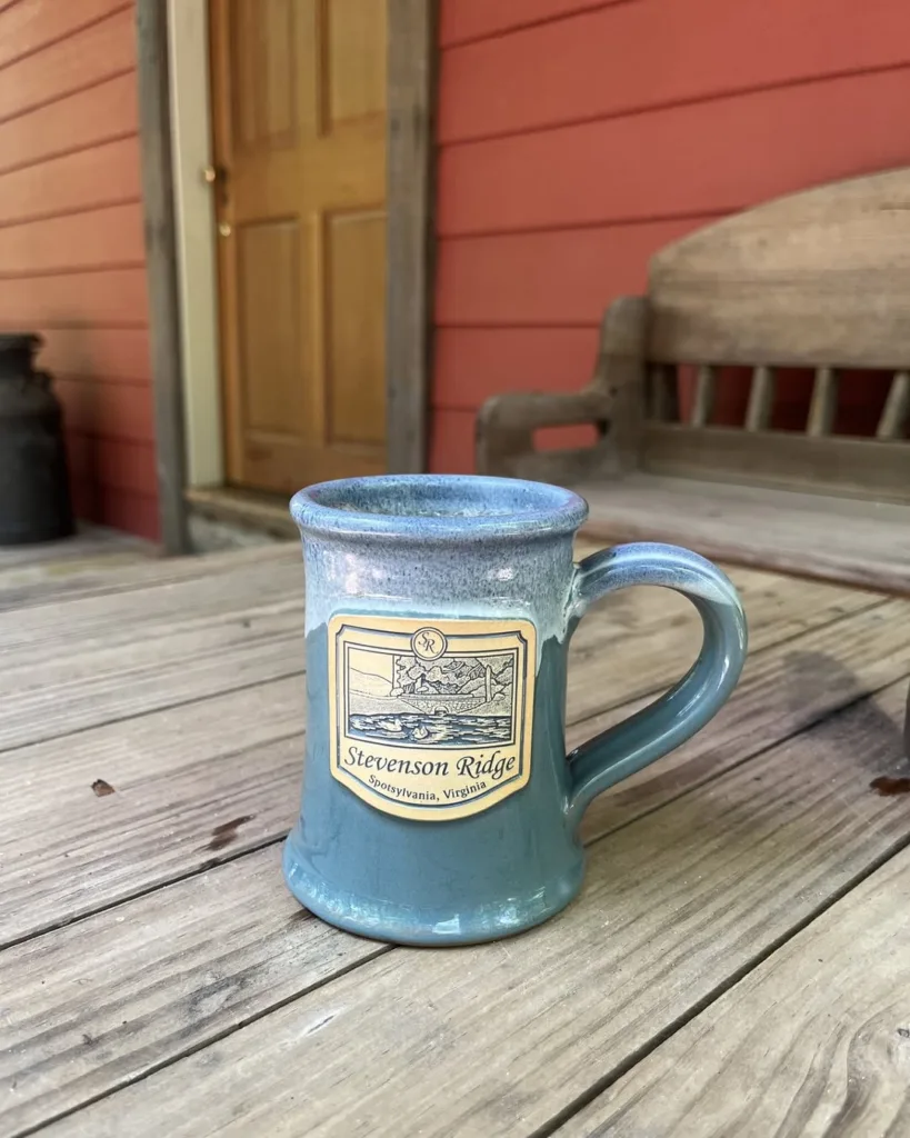 Close-up of a blue and gray ceramic mug branded with the Stevenson Ridge logo and location (Spotsylvania, Virginia), resting on the wooden porch of the Adrian Cabin, promoting historic cabin lodging.