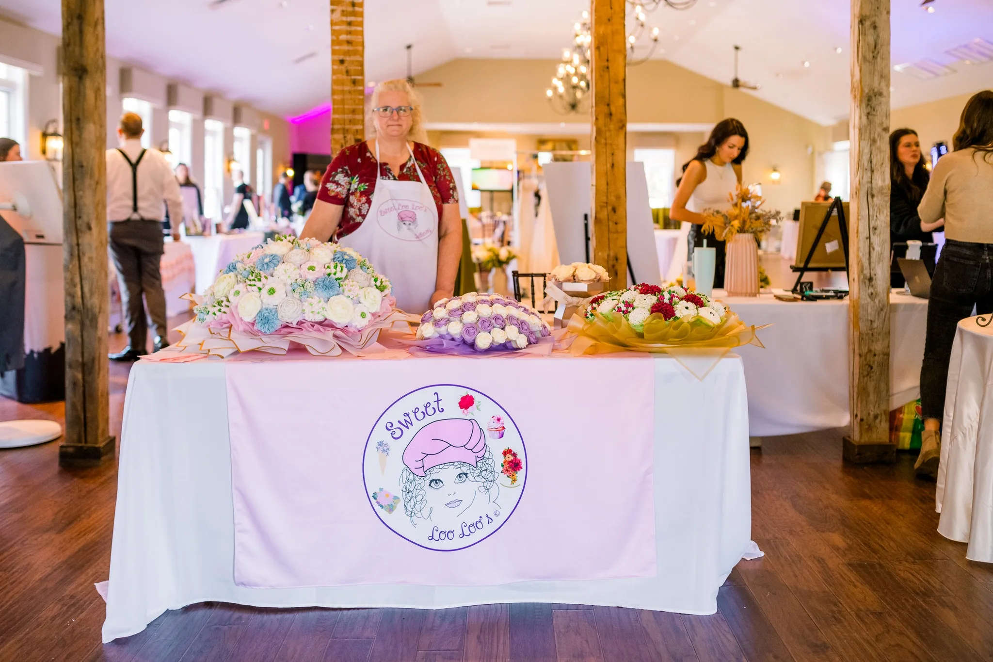 Female vendor from Sweet Loo Loo's standing behind a table displaying colorful and uniquely arranged cupcake bouquets inside the Stevenson Ridge event space with rustic wood beams.