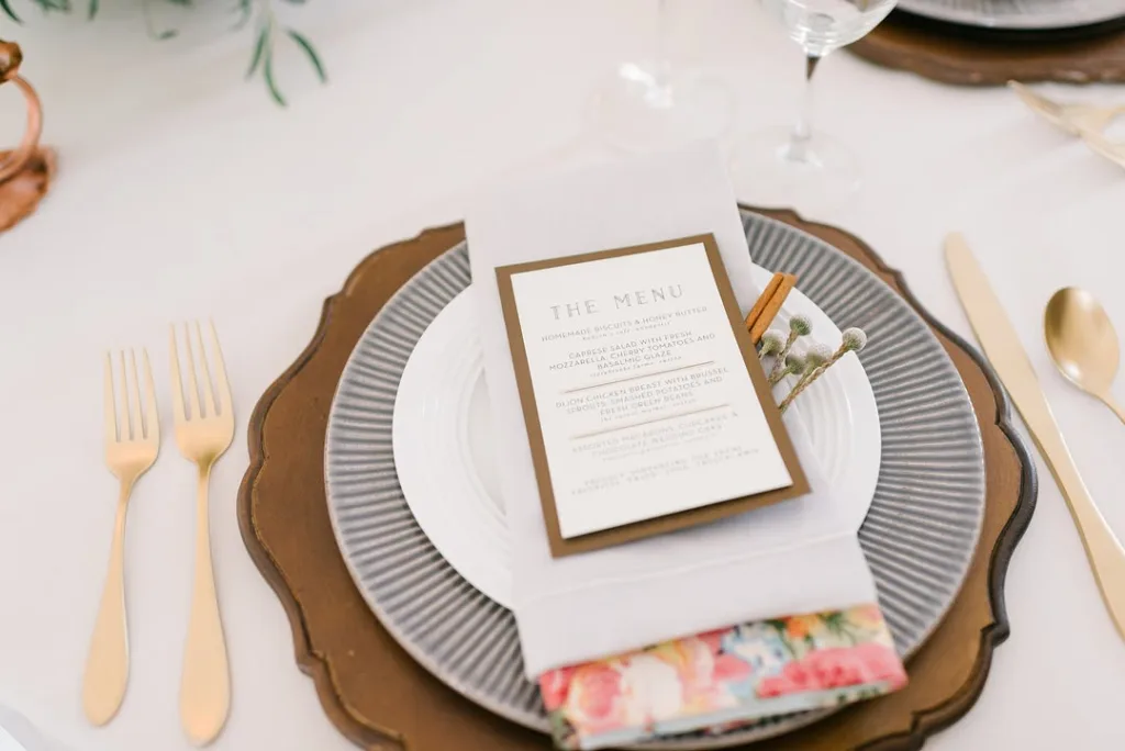 Detailed place setting featuring a custom printed menu card on a white napkin, layered plates (gray and wood charger), and gold forks and knife, showcasing formal dining details for a Stevenson Ridge wedding.