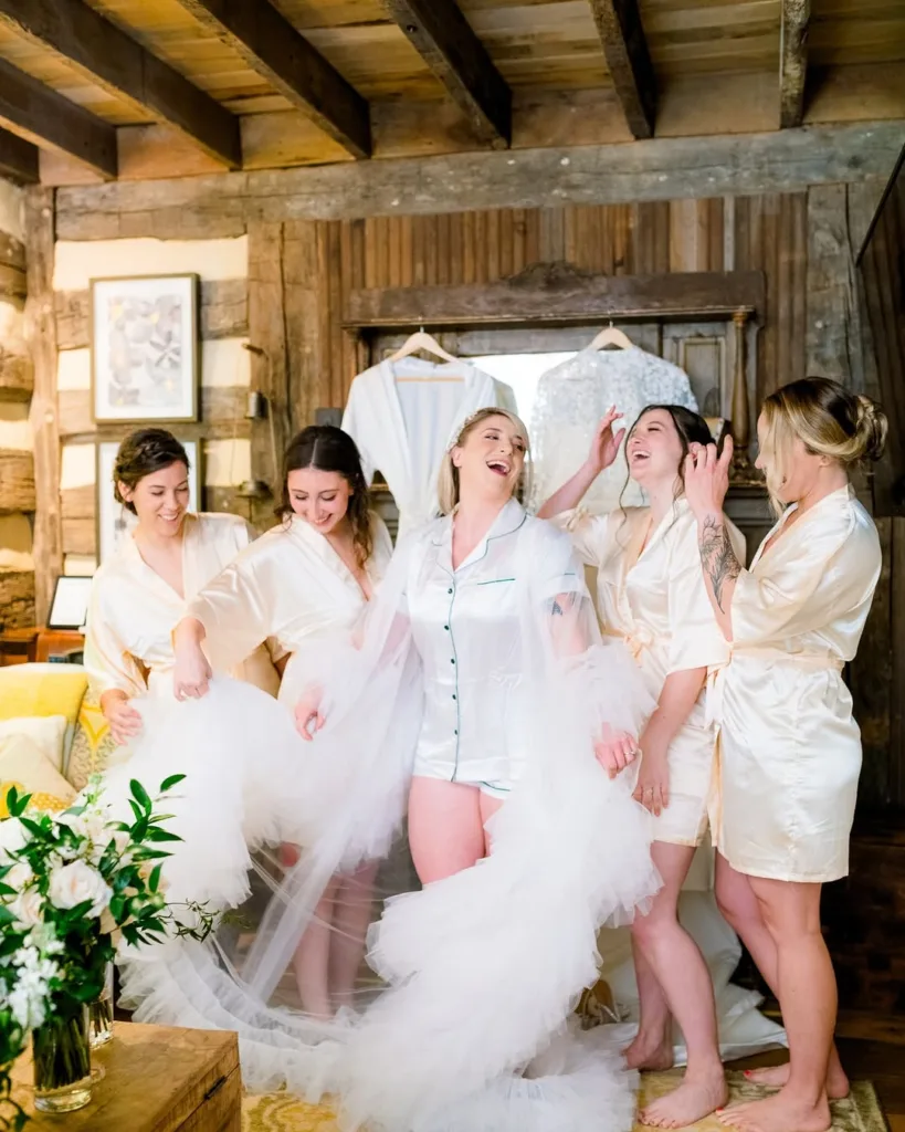 The bride and four bridesmaids laughing and playing with the bride's tulle veil while wearing matching satin robes and pajamas inside a rustic Stevenson Ridge log cabin, capturing fun Spotsylvania getting-ready photos.