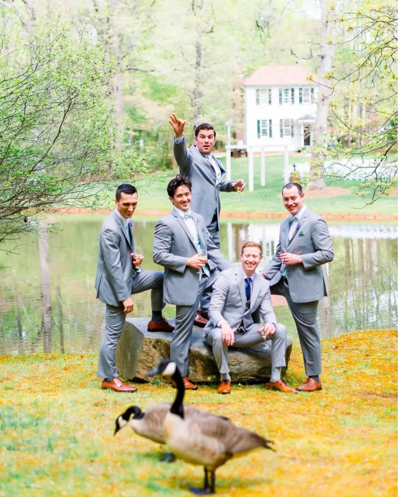 Five groomsmen in light gray suits posing on a large rock next to the lake at Stevenson Ridge, with a Canada goose in the foreground and a white historic house in the background, perfect for Spotsylvania wedding photography.