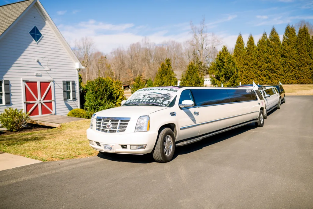 A white Cadillac stretch limousine from Fredericksburg Limousine parked on the asphalt in front of a gray building with red barn doors at Stevenson Ridge, showcasing Spotsylvania wedding transportation.