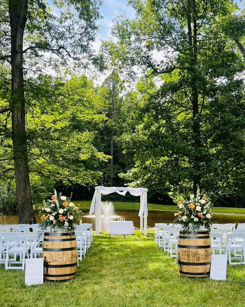 Outdoor wedding ceremony site at Stevenson Ridge with white chairs, a white arbor, floral arrangements on wine barrels, and a scenic pond with a fountain in the background, set amidst lush green trees in Spotsylvania.