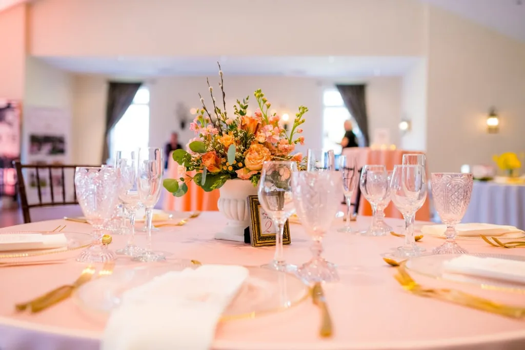 Wedding reception table set with a peach tablecloth, clear and textured glassware, gold flatware, and a coral and peach floral centerpiece, showing the brightly lit interior of the Stevenson Ridge Lodge in Spotsylvania.