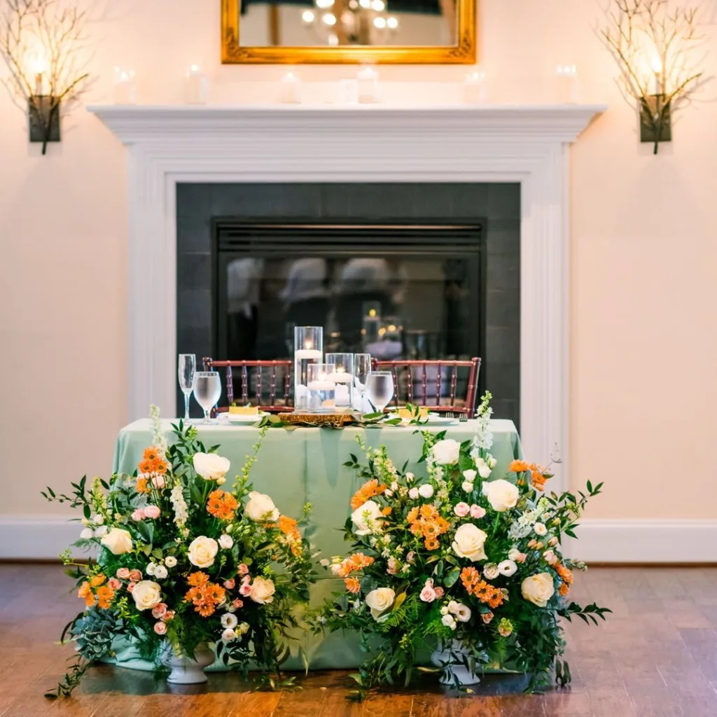 A dramatic sweetheart table covered in a sage green linen, framed by two large floor arrangements of orange, white, and peach flowers, set in front of a working fireplace in the Stevenson Ridge Lodge.