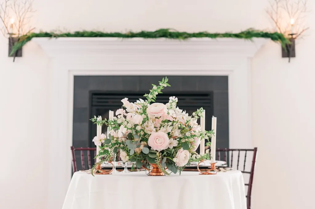 Close-up of a small wedding table set for two (sweetheart table) with a large blush pink floral centerpiece, tall candles, and copper accents, set against the white mantel and black fireplace in the Stevenson Ridge Lodge.
