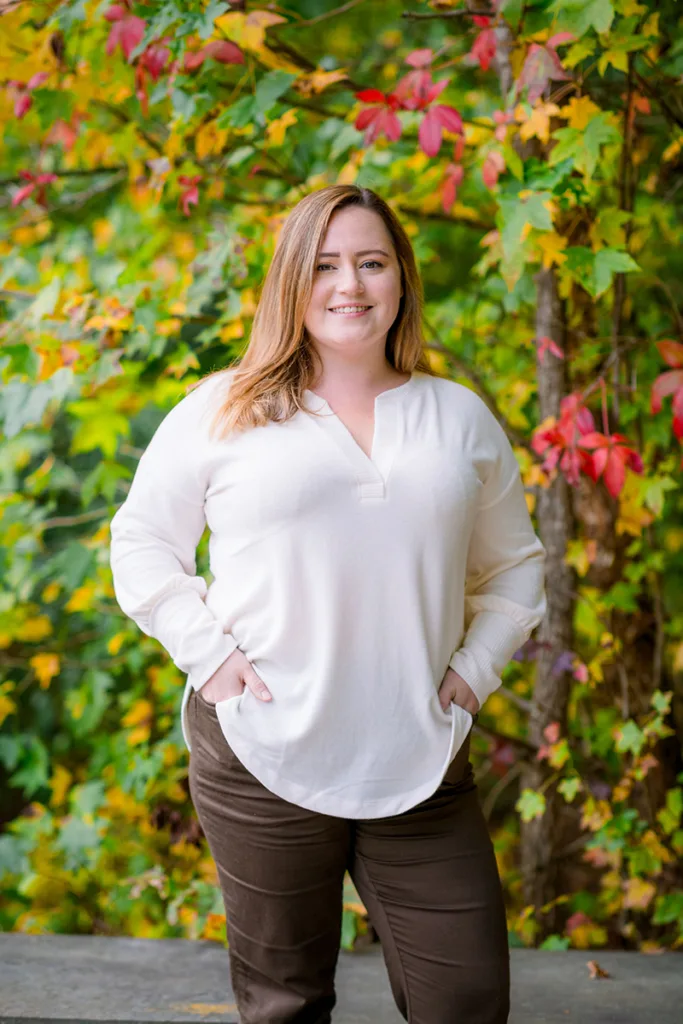 Professional portrait of Taylor Buel, the Stevenson Ridge Event Coordinator, smiling in a cream sweater against a backdrop of vibrant red and yellow autumn foliage, for Spotsylvania wedding planning services.