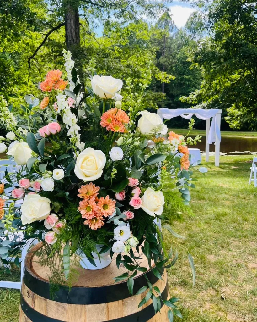 Close-up of a rustic wine barrel topped with a lush wedding floral arrangement featuring white roses, peach dahlias, pink carnations, and greenery, with an outdoor ceremony setup blurred in the background at Stevenson Ridge.