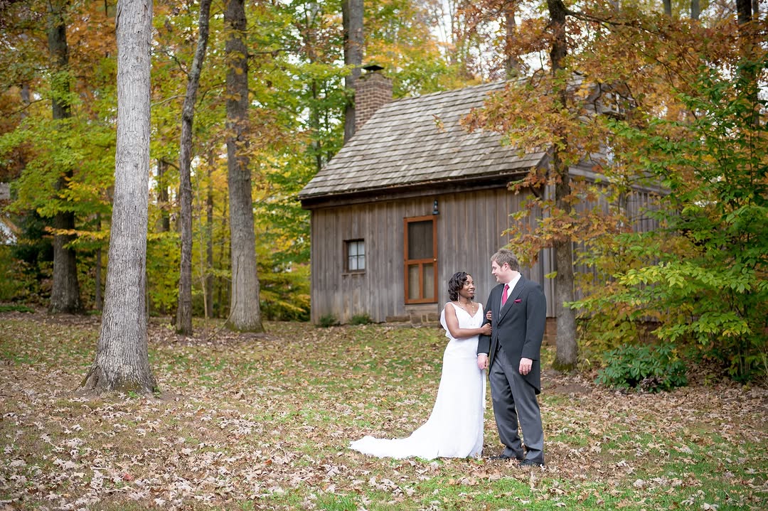 Portrait of a smiling bride and groom standing in front of the rustic 18th-century cabin at Stevenson Ridge, surrounded by colorful autumn foliage and fallen leaves, ideal for a Spotsylvania elopement.