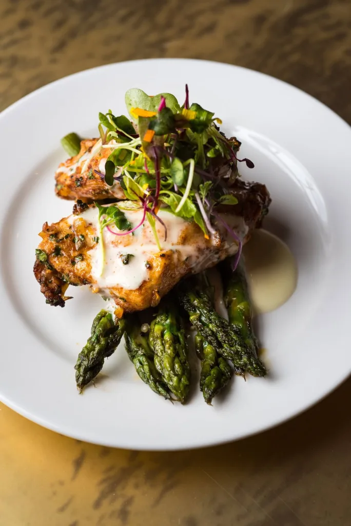 Top-down view of a beautifully plated dish featuring two pieces of herb-crusted chicken, roasted asparagus, a creamy sauce, and a fresh garnish of microgreens, showcasing Stevenson Ridge wedding catering in Fredericksburg.