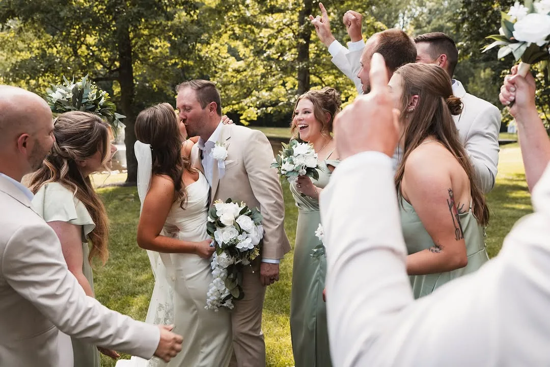 Candid photo of the newly married couple kissing, surrounded by their laughing wedding party in tan and sage green attire, holding white rose bouquets in a sunny outdoor wooded area at Stevenson Ridge.