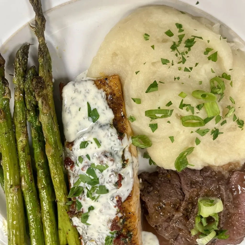 Close-up of a plated dinner featuring a piece of pan-seared salmon topped with creamy sauce, a slice of beef tenderloin (surf and turf), mashed potatoes, and roasted asparagus, showcasing Stevenson Ridge wedding catering.