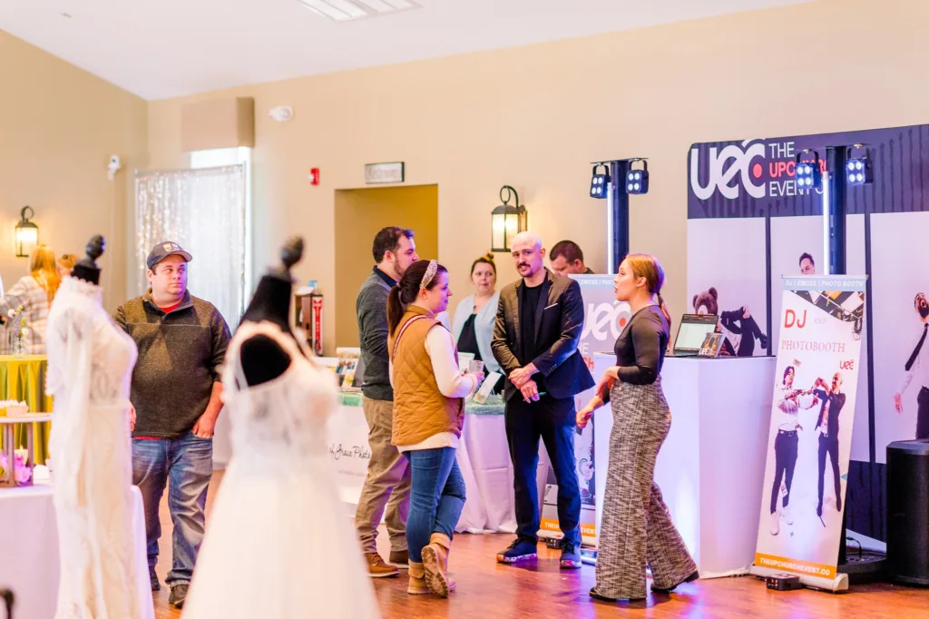Engaged couples interacting with a male DJ vendor from The Upchurch Event Company, who stands in front of his branded setup, with wedding dresses in the foreground at a Stevenson Ridge showcase.