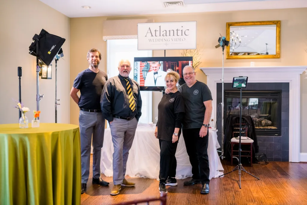Four members of the Atlantic Wedding Video team posing with their equipment, including a display monitor, lights, and branding, in a bright room at a Stevenson Ridge vendor show in Fredericksburg, Virginia.
