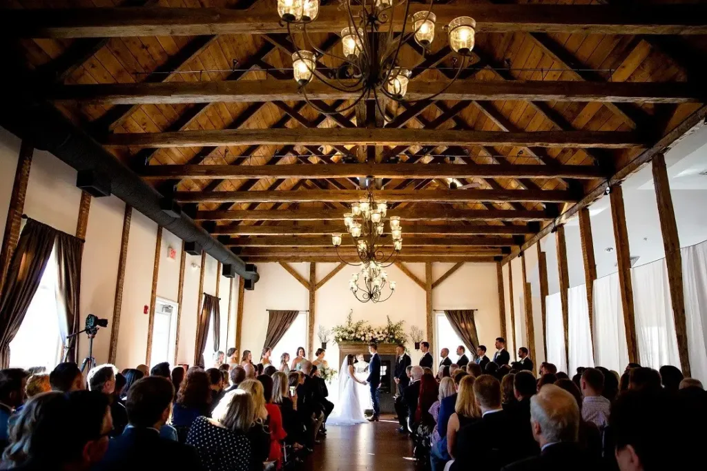 A bride and groom exchanging vows in front of a stone fireplace during an indoor wedding ceremony inside The Lodge at Stevenson Ridge, featuring heavy exposed wooden beams and large chandeliers.